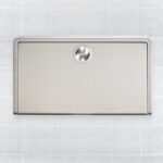 Stainless steel wall-mounted baby changing station on tiled wall, closed panel.