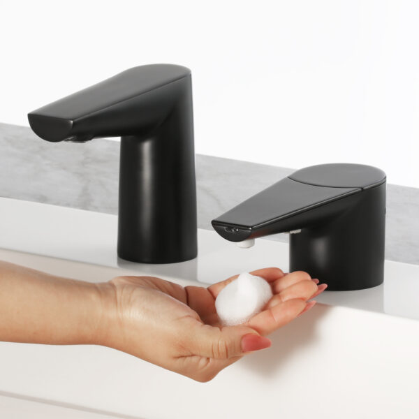 Hand under black automatic soap dispenser, foam dispensing, another dispenser in background.