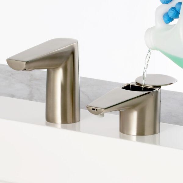 Person with blue gloves pours liquid into metallic soap dispenser by faucet on countertop.