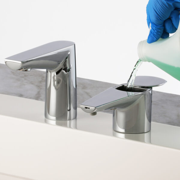 Gloved hand pours green liquid into metallic soap dispenser next to faucet on countertop.