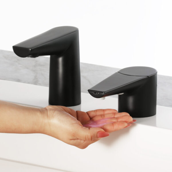 Person using black automatic soap dispenser dispensing pink soap. Another dispenser nearby.