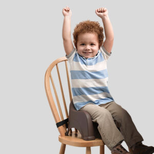 Child in booster seat on wooden chair, arms raised, wearing striped shirt and khaki pants.