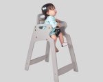 Toddler in light blue outfit seated in gray high chair with safety strap and blue hair bow.